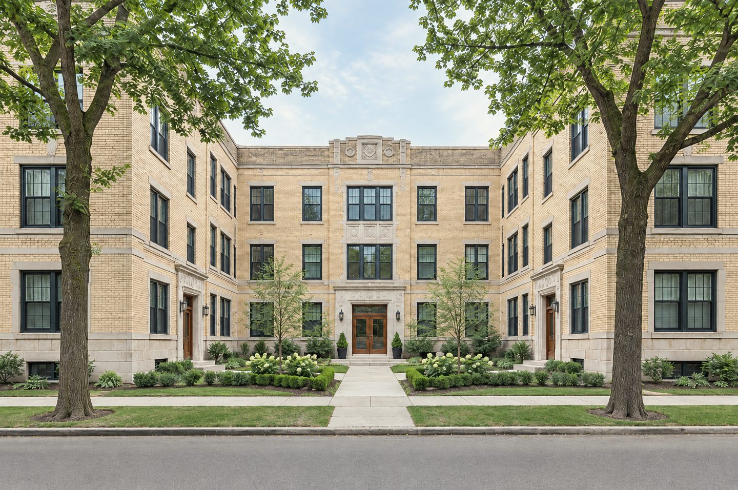 A renovated three-storey 1920s courtyard apartment building in cream brick with limestone trim, black-framed windows and a landscaped front courtyard on a tree-lined street.