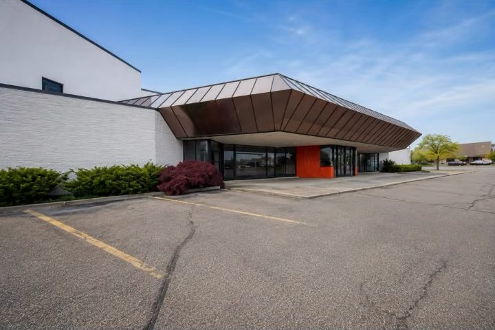 The building at 6750 Orchard Lake Road, West Bloomfield: white brick walls and an angular copper-toned canopy over a glass and orange-panelled entrance, seen across its parking lot on a clear day.