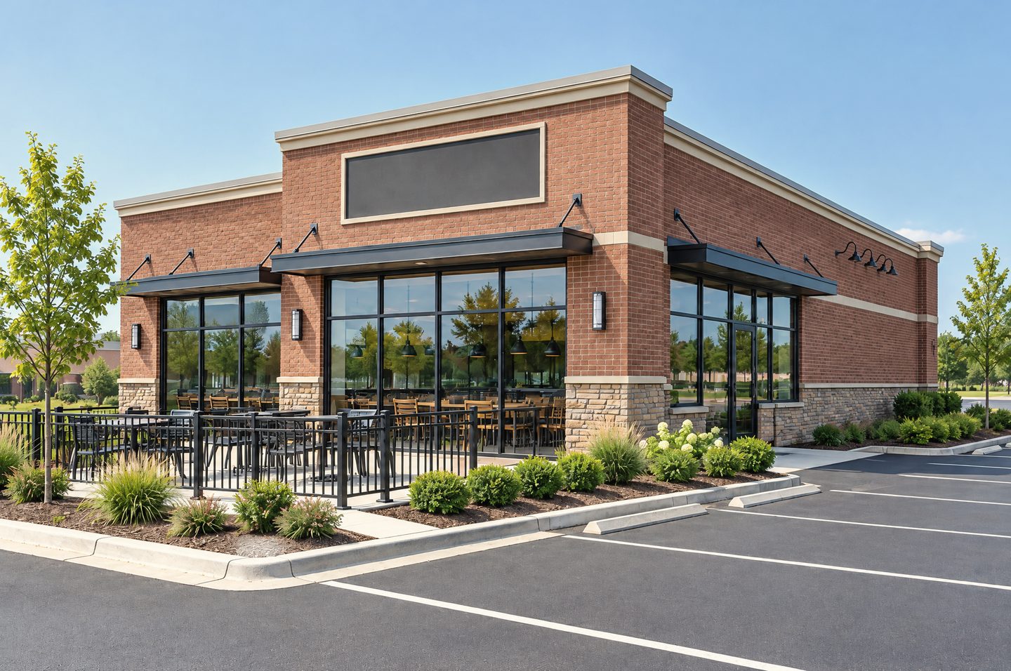 A standalone single-storey restaurant and bar in red-brown brick with a stone base, tall storefront windows, a railed front patio with tables and a freshly striped parking lot on a sunny day.