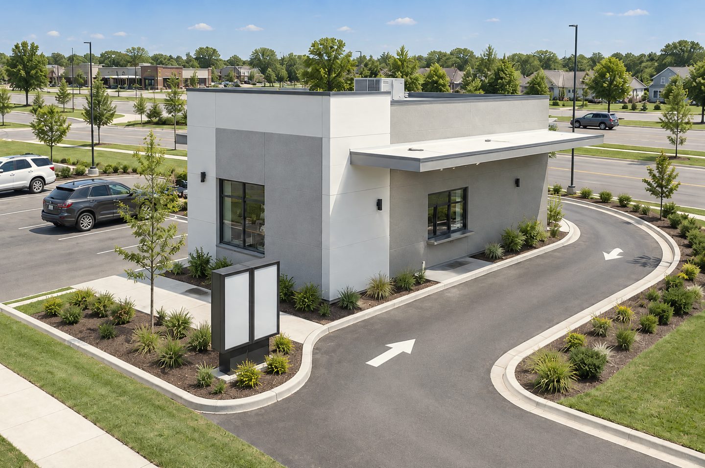 Elevated daylight view of a small standalone quick-service restaurant on its own pad: grey and white panels, a canopy over the pick-up window and a curved drive-thru lane with painted arrows beside an arterial road.